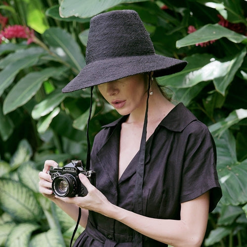 Black MEG jute straw hat with tall crown and medium brim worn by woman holding camera outdoors