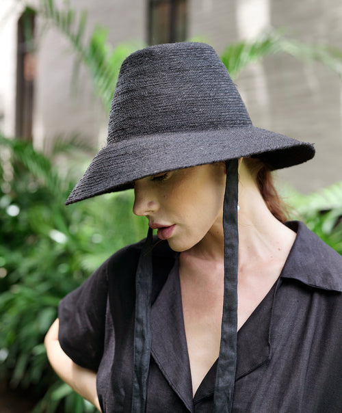 Black MEG jute straw hat with tall crown and medium brim, featuring natural jute material and linen lining, worn with neck straps.