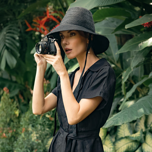 Woman wearing black MEG jute straw hat with tall crown and medium brim holding a camera outdoors